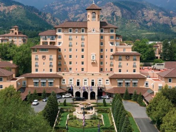 A large, elegant hotel with a central tower, surrounded by trees and mountains in the background, with a landscaped garden in front.