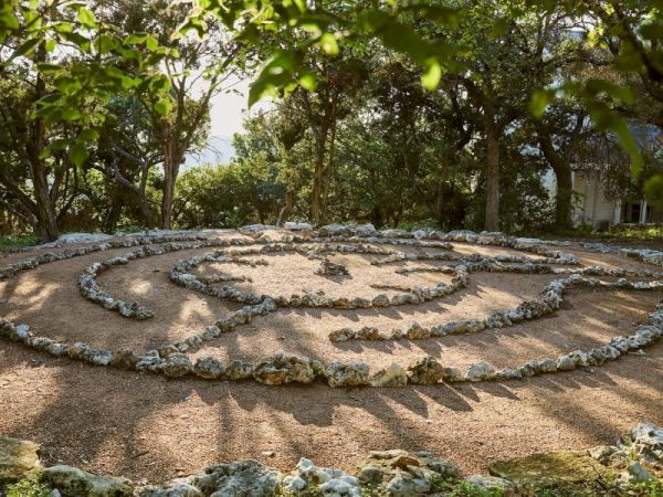 The image shows a stone labyrinth in a garden setting surrounded by trees, with sunlight casting shadows on the path.