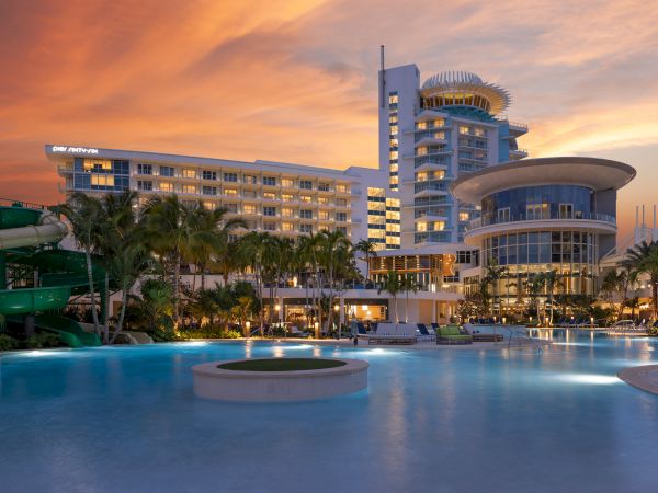 A luxurious resort with a swimming pool, palm trees, and a sunset sky, featuring modern architecture and a serene ambiance.