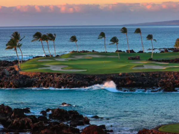 A coastal golf course with palm trees, rocky shore, and ocean waves under a colorful sky during sunset.