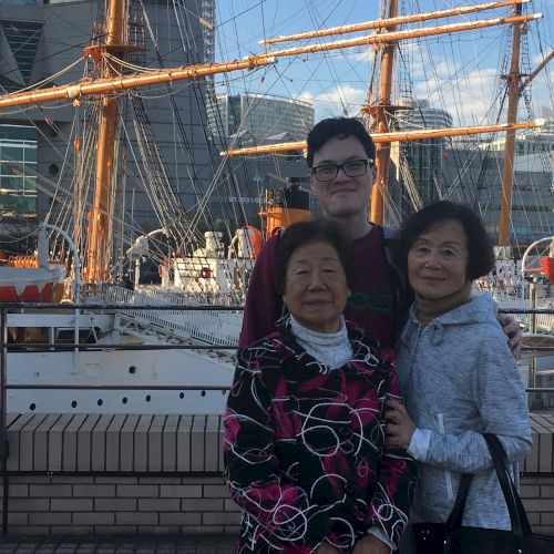 Three people are posing in front of a vintage sailing ship docked at a harbor, with buildings in the background.