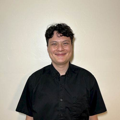 A person is standing against a plain wall, smiling, wearing a black shirt.