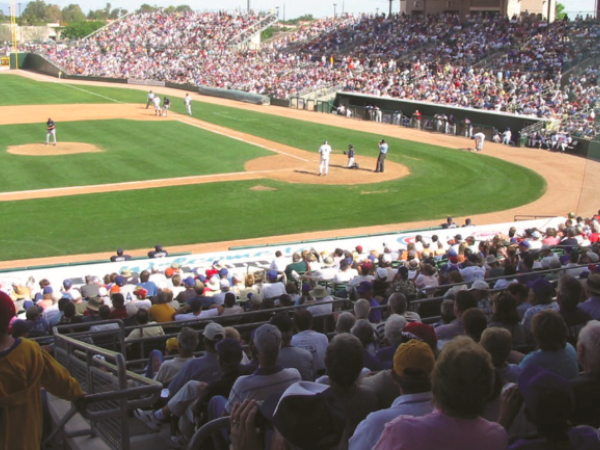 A crowded baseball stadium with fans in the stands, people on the field, and a game in action on a sunny day.
