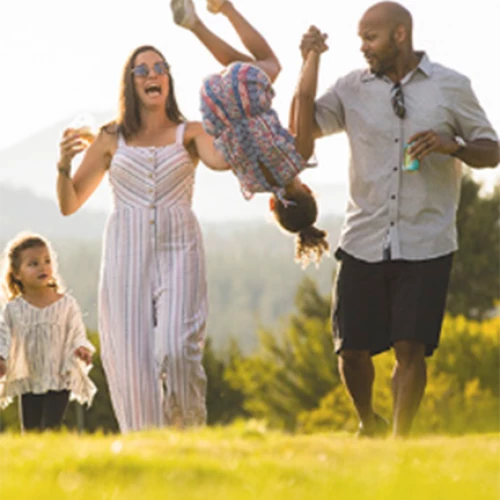 A family of four enjoys the outdoors, with the parents swinging one child by her arms while the other child walks beside them.