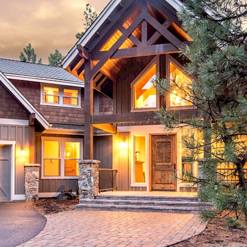 A large, illuminated house with wood and stone accents, multiple windows, and a double garage is set amidst tall trees in the evening.