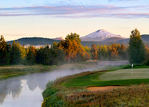 A serene golf course beside a calm river at sunrise with mist and a backdrop of forested mountains, including one snow-capped peak, is depicted.