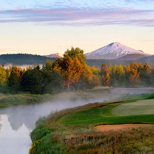 A serene golf course beside a calm river at sunrise with mist and a backdrop of forested mountains, including one snow-capped peak, is depicted.