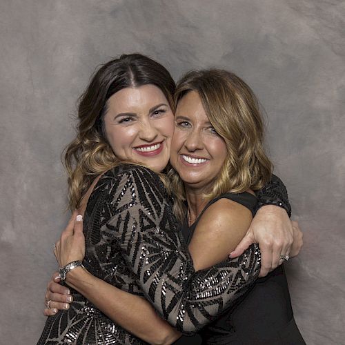 Two women are happily embracing each other while smiling at the camera. Both of them are dressed in formal attire, standing in front of a neutral backdrop.