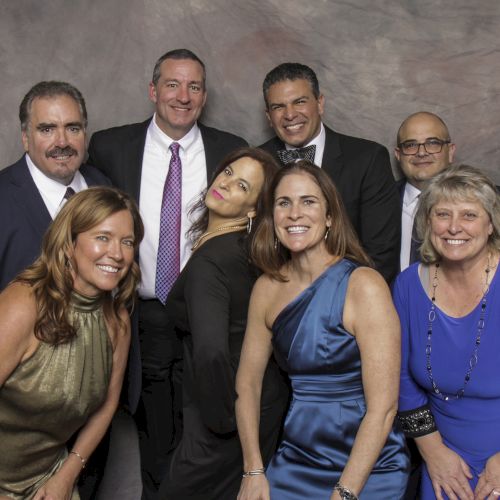 A group of seven people are dressed in formal attire, posing together and smiling against a neutral backdrop.