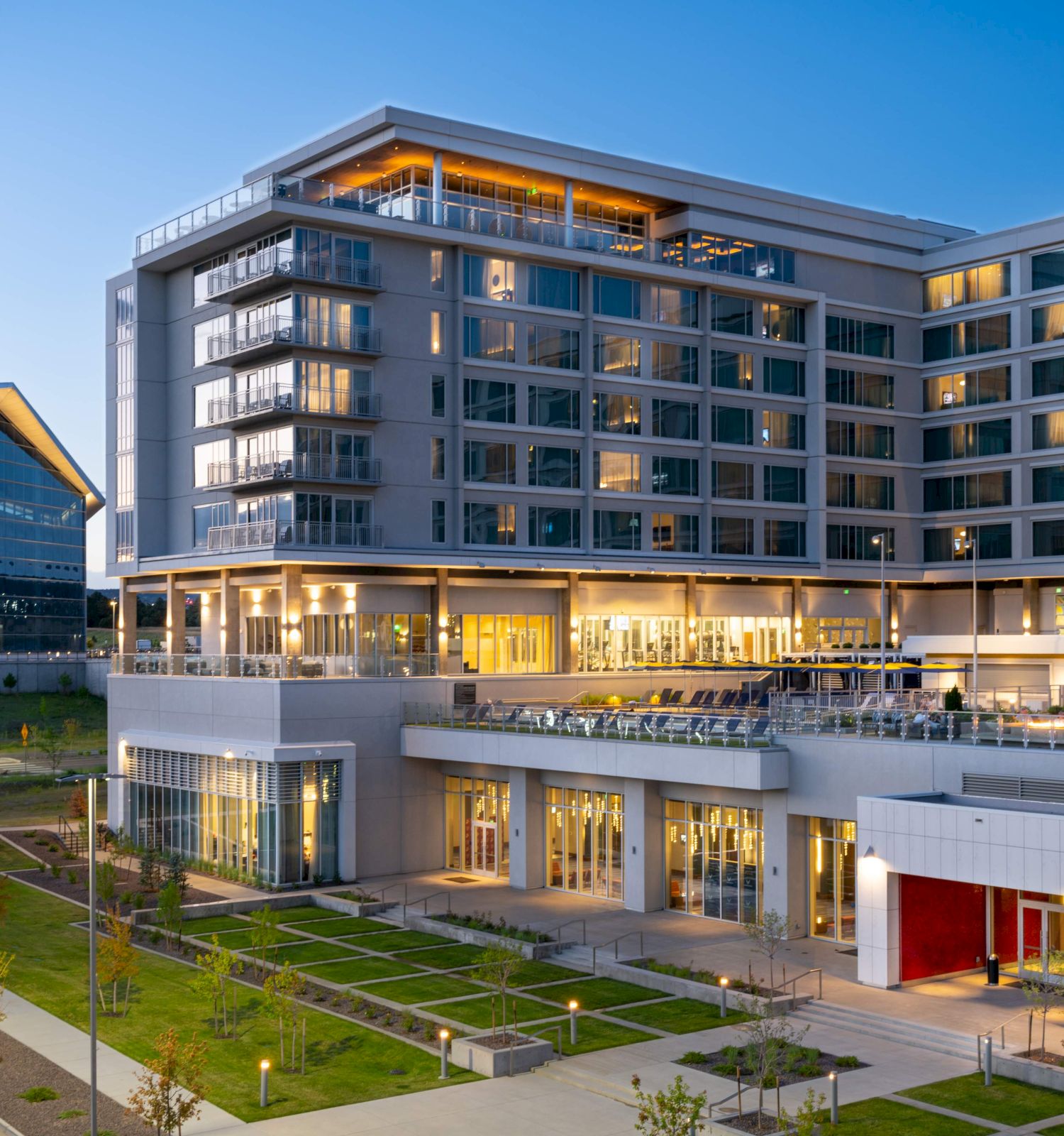A modern multi-story office/campus building with balconies, large windows, and warm lights at dusk.