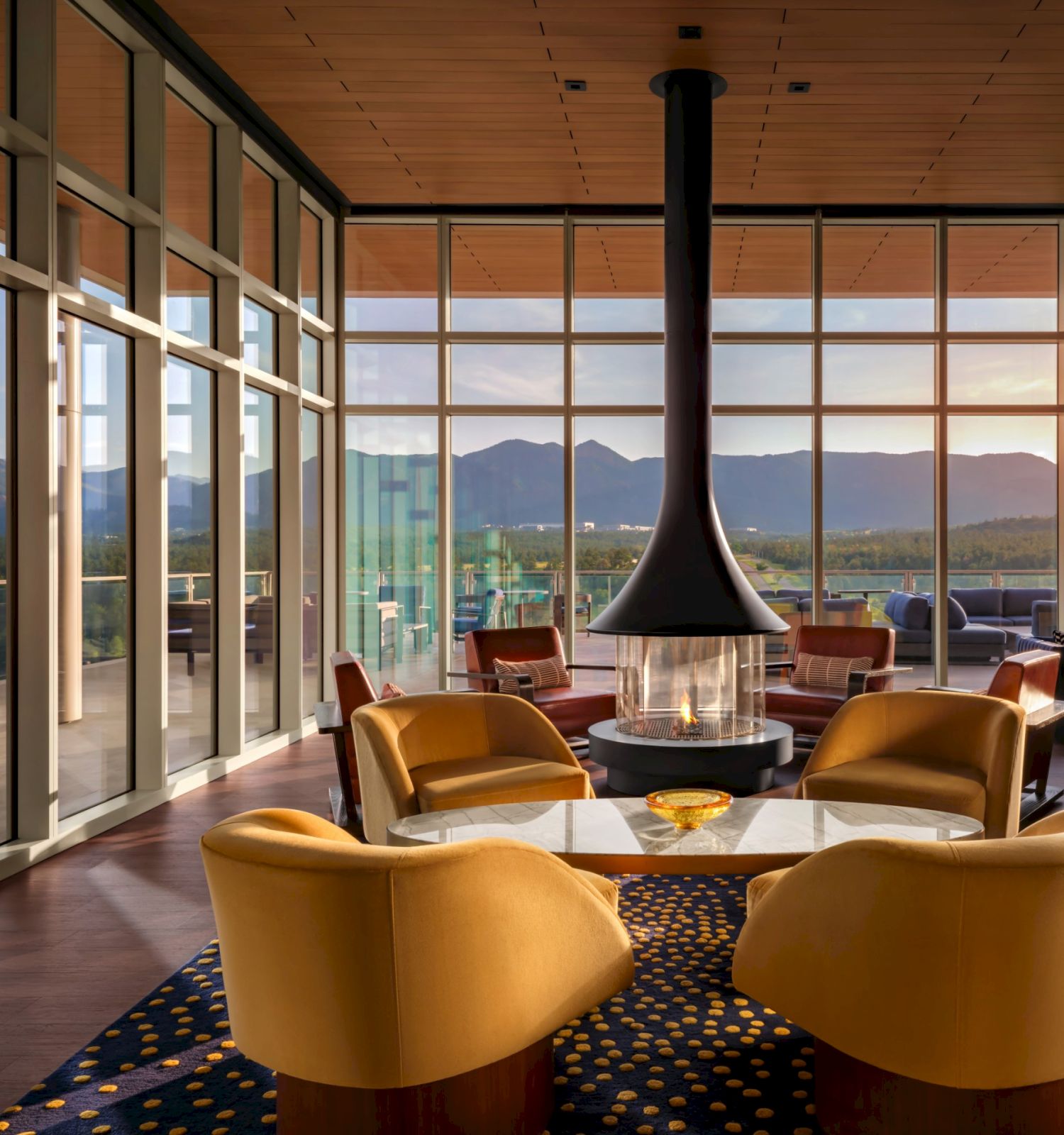 A modern lounge with large windows, mountain sunset views, stylish chairs, and a central fireplace/heat lamp.