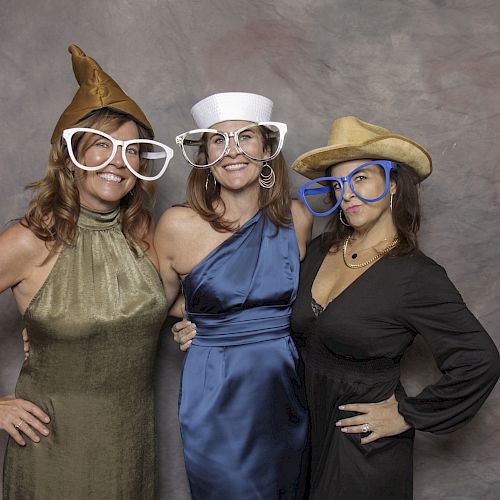 Three women are posing together, each wearing different hats and oversized glasses, while standing against a neutral backdrop.