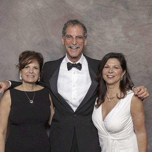 Three people, two women and one man, are posing and smiling in front of a neutral backdrop. They're dressed formally.