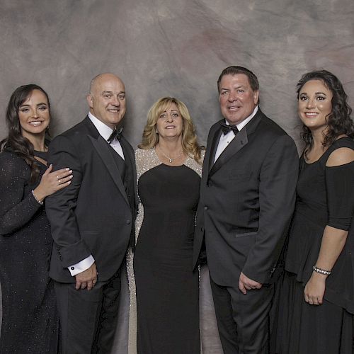 A group of five people, dressed in formal attire, stand together against a neutral, textured backdrop, smiling for the camera.