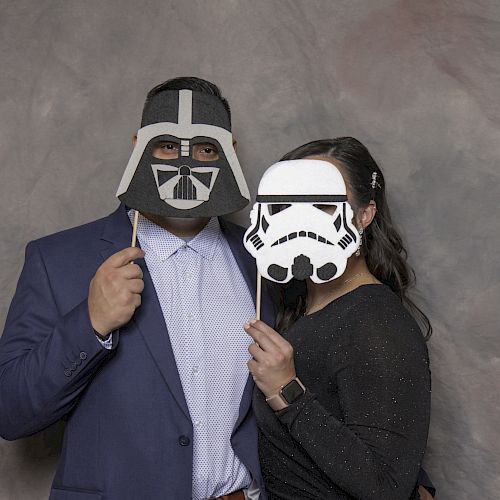 Two people dressed formally are holding masks resembling characters from Star Wars, standing in front of a grey backdrop.