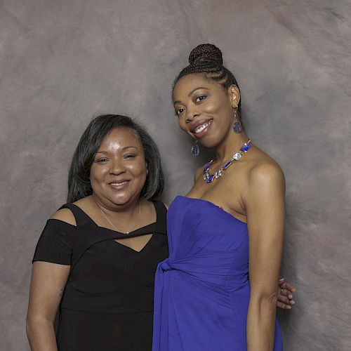 Two women are posing for a photo together, one in a black dress and the other in a blue dress, smiling against a neutral backdrop.