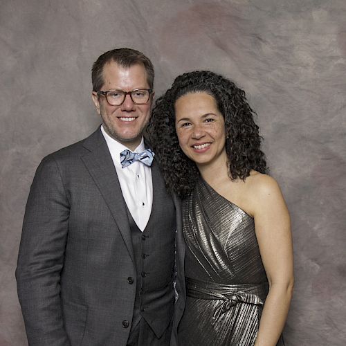 A man in a gray suit with a bow tie and a woman in a metallic dress are posing against a textured gray backdrop.