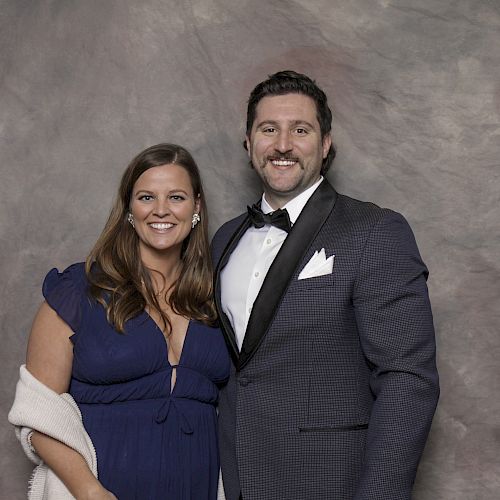 A couple poses for a formal photo; the man wears a tuxedo, and the woman is in a blue dress, against a grey backdrop.