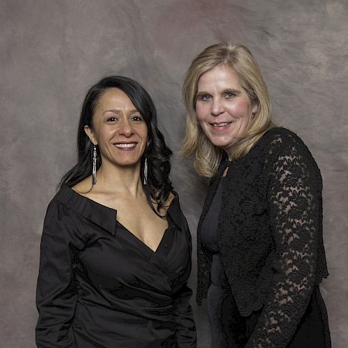 Two women are standing and smiling in front of a plain backdrop, both dressed in black outfits.