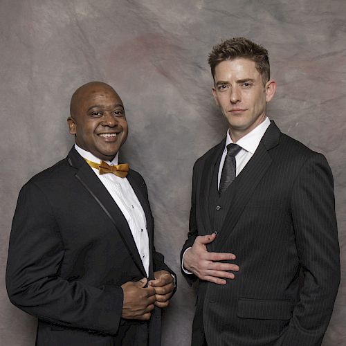 Two men in suits posing for a photo against a textured backdrop, one smiling, the other with a neutral expression.