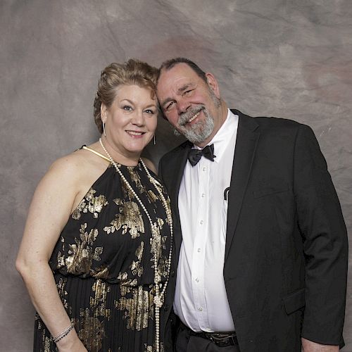 A man and woman are dressed in formal attire, standing together against a gray backdrop. The man is in a black suit; the woman wears a patterned dress.