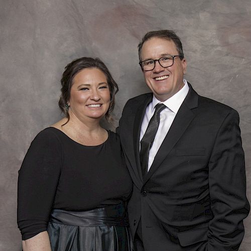 A man and a woman are posing together, both dressed formally in black attire, with a gray and textured background behind them.