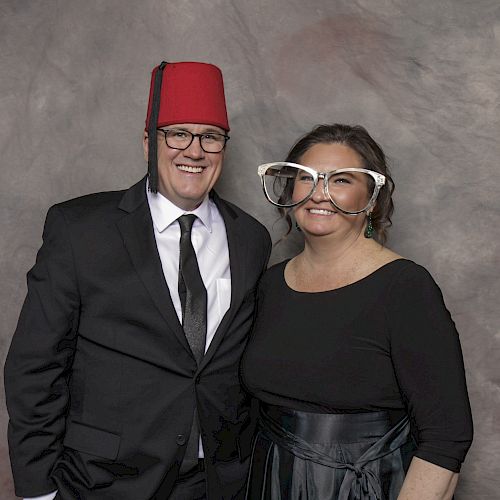 A man in a suit and fez hat stands next to a woman in a black dress and oversized glasses; both are smiling in front of a gray backdrop.