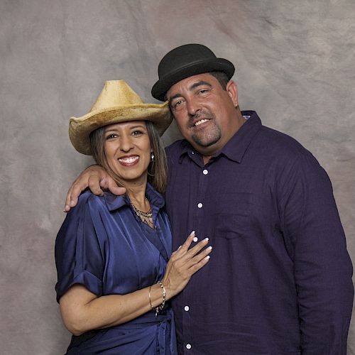 A woman wearing a cowboy hat and a man in a bowler hat are standing together, smiling at the camera, with his arm around her shoulder.