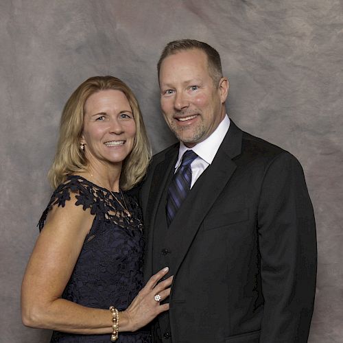 A couple dressed formally, the woman in a black dress and the man in a black suit, pose against a light gray backdrop, both smiling at the camera.