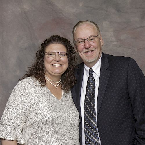 A smiling couple poses in front of a gray textured backdrop. The woman wears a light-colored outfit; the man wears a dark suit and tie.