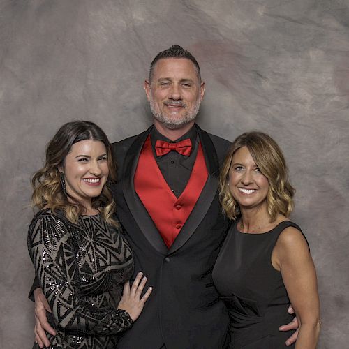 A man in a suit with a red vest and bow tie stands between two women, all smiling, against a gray backdrop.