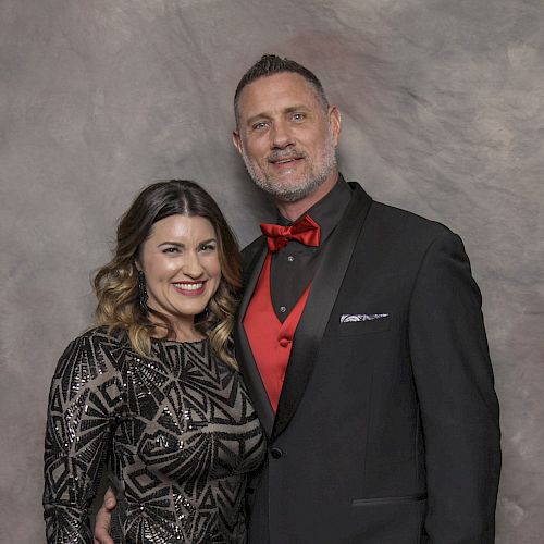 A man in a black suit with a red bow tie and vest stands next to a woman in a patterned dress, both smiling.