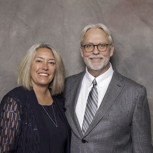 A man and woman are standing together, smiling at the camera. The man is wearing a gray suit, and the woman is wearing a dark dress.