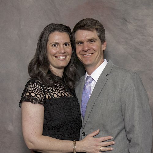 A smiling couple poses together, the woman in a black dress, and the man in a gray suit with a light purple tie, against a plain backdrop.