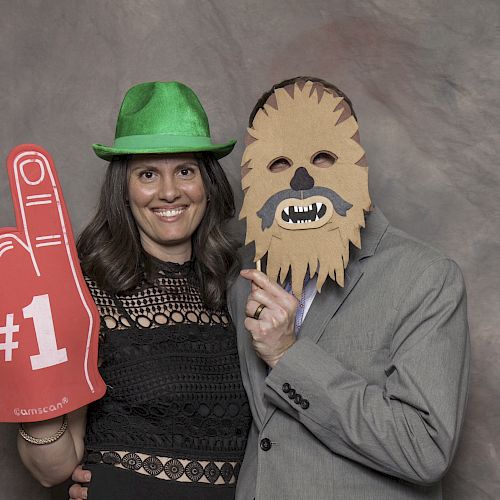 A woman in a green hat stands next to a person in a suit wearing a mask and holding a foam finger with 