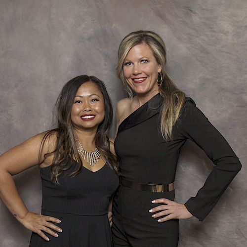 Two women are standing close to each other, smiling and posing for the camera in front of a neutral backdrop. Both are dressed in black outfits.