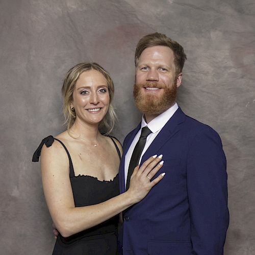 A couple poses for a portrait in front of a studio backdrop; the woman wears a black dress, and the man is in a blue suit.