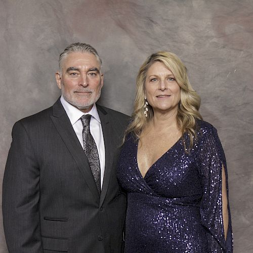 A man in a suit and a woman in a sparkling purple dress are posing in front of a neutral backdrop.