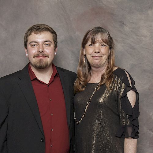 A man and a woman are standing together, smiling, in front of a gray and brown textured backdrop. The man is in a suit, and the woman is in a dress.