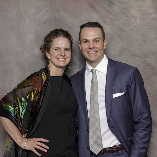 A woman in a black dress and a man in a blue suit, white shirt, and gray tie are standing in front of a marbled backdrop, smiling at the camera.