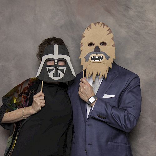 Two people are posing for a photo while holding up masks of Darth Vader and Chewbacca, standing in front of a neutral backdrop.