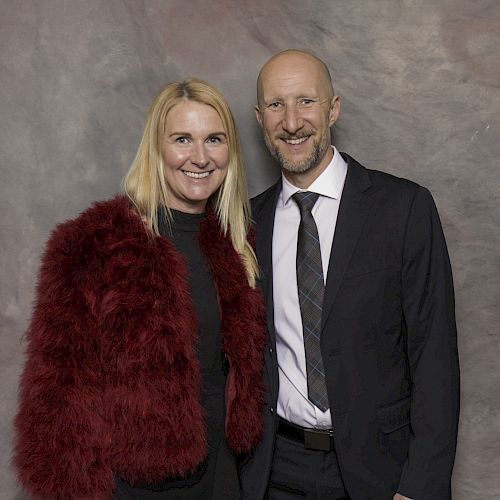 A man and a woman are smiling at the camera. The woman wears a red fur jacket, and the man is in a suit with a tie.