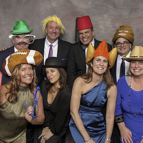 A group of people in formal attire, wearing funny hats and props, posing for a playful photo against a neutral backdrop.