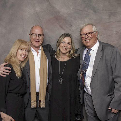 Four adults dressed in formal attire are standing together, smiling at the camera against a gray backdrop.