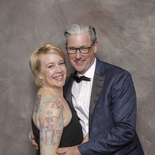 A smiling couple, the woman with blonde hair and tattoos, and the man in glasses and a suit with a bow tie, against a gray backdrop.