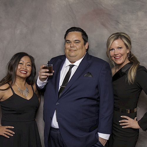 Three people are posing together, dressed in formal attire; the man is holding a drink and standing between two women, against a neutral backdrop.