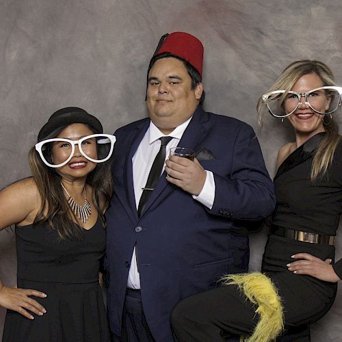 Three people pose in costumes with oversized glasses, one wearing a fez. The setting appears playful with a neutral backdrop.